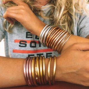 A person wearing a gray t-shirt with red and white lettering holding their curly blonde hair up, showingcasing multiple metallic bangles on their wrists.
