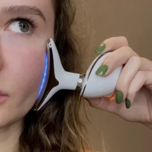 Uma mulher segurando um dispositivo de massagem facial próximo à bochecha, com esmalte verde perceptível nas unhas e uma visão parcial do rosto com foco em um olho.